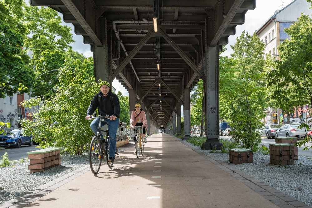 Ein Bild zeigt zwei Radfahrer, die unter einem Bahndamm entlang eines begrünten Radwegs fahren. Das Foto zeigt einen sonnigen Tag mit zwei Personen, die mit Fahrrädern unter einem Bahndamm fahren. Der Mann fährt vorne und trägt einen Helm und dunkle Kleidung. Die Frau hinter ihm trägt einen hellen Pullover und einen Helm. Der Radweg ist gepflastert und von Grünflächen, Bänken und kleinen Mauerabschnitten sowie Bäumen gesäumt. Der graue Bahndamm über dem Radweg ist mit Stahlträgern und Beleuchtung ausgestattet, und er erstreckt sich weit in die Ferne. Im Hintergrund sind Wohnhäuser und Straßen mit parkenden Autos zu sehen. Die Atmosphäre ist ruhig und friedlich, die Stimmung entspannt und sonnig. Das Bild vermittelt ein Gefühl von städtischer Natur und nachhaltiger Mobilität.