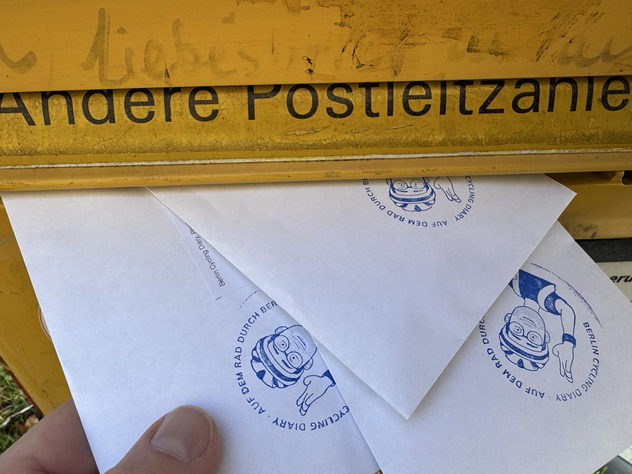 Ein Finger schiebt Briefe mit einem Fahrradmotiv in einen gelben Briefkasten. Das Bild zeigt einen Ausschnitt eines gelben Briefkastens, in den mehrere weiße Briefumschläge gesteckt werden. Der Briefkasten trägt die Aufschrift "Andere Postleitzanie", und darüber ist in dunkler Schrift das Wort "Liebes" zu erkennen. Die Briefumschläge sind mit einem blauen Aufdruck versehen, der ein stilisiertes Gesicht mit Fahrradhelm und den Schriftzug "BERLIN CYCLING DIARY. AUF DEM RAD DURCH BERLIN" zeigt. Ein menschlicher Finger ist im Vordergrund zu sehen, der einen der Briefumschläge hält. Die Stimmung des Bildes ist alltäglich und pragmatisch, da es eine gewöhnliche Handlung des Postversands darstellt.