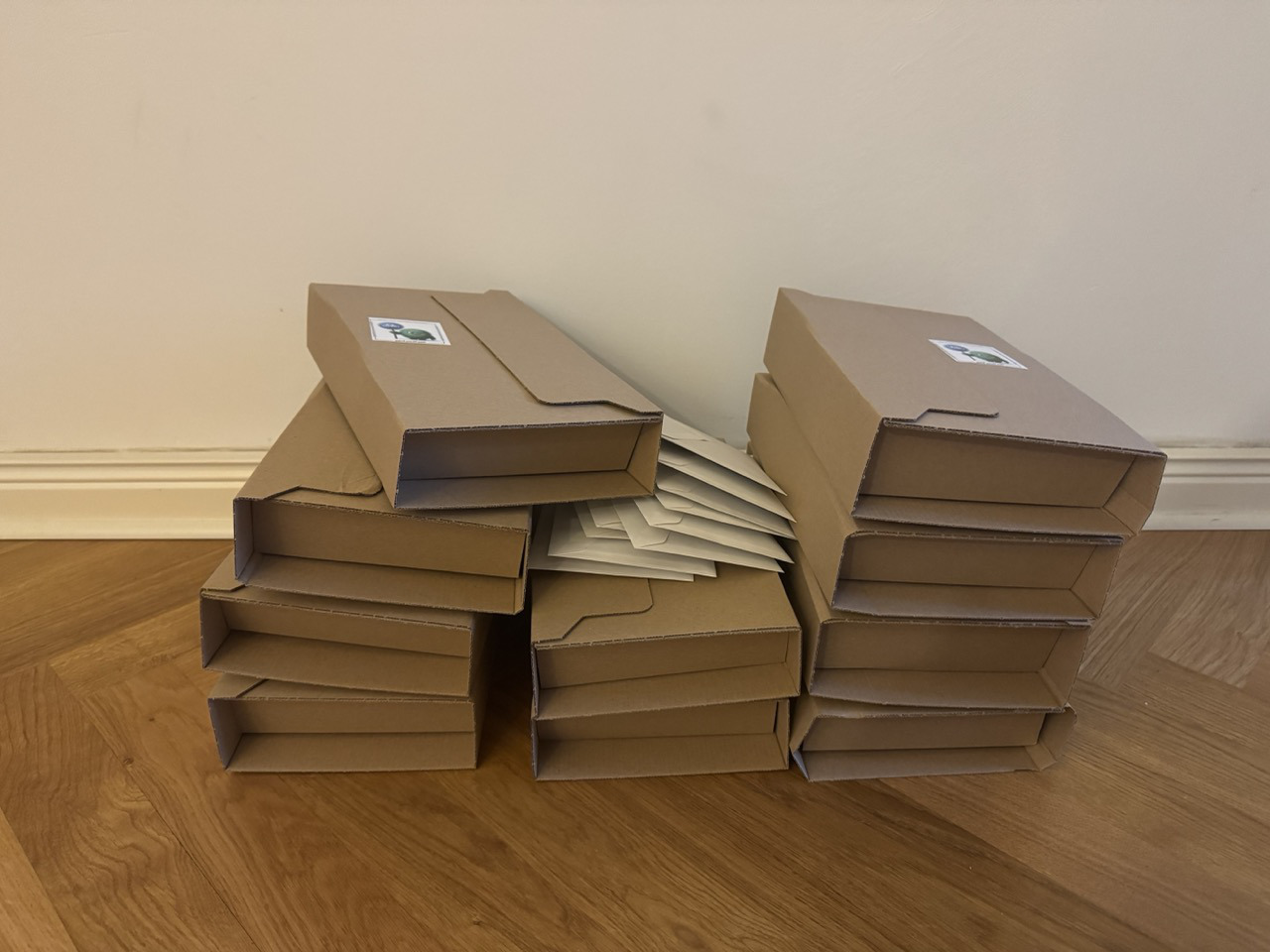 Das Bild zeigt zwei Stapel von braunen Kartons auf einem Holzboden vor einer weißen Wand. Zwischen den Kartons liegen einige flache, braune Umschläge. Auf der Oberseite der Kartons ist jeweils ein kleiner Aufkleber zu sehen.